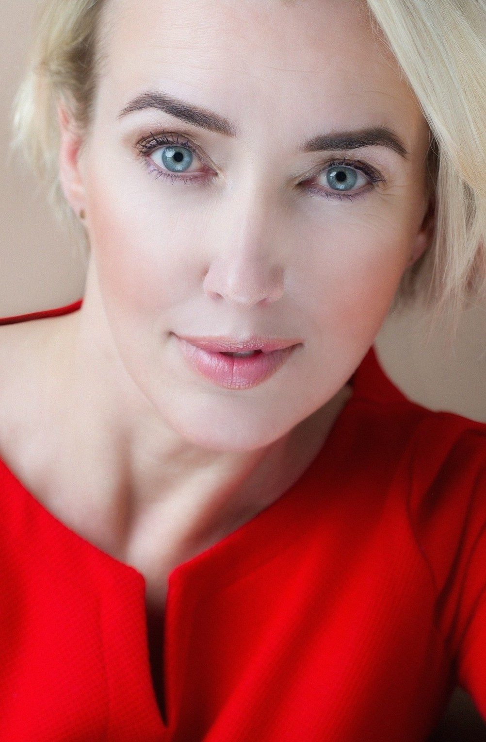 Nahes Beautyportrait einer blonden Frau mit blauen Augen in roter Bluse vor beigem Hintergrund