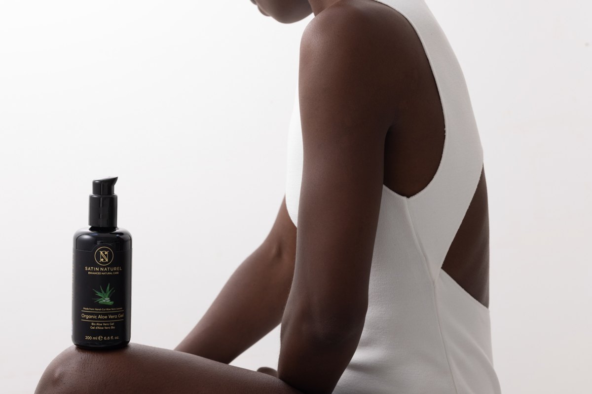 Dunkle Aloe-Vera-Gel-Flasche von Satin Naturel auf dem Knie einer sitzenden Frau in weißem Body