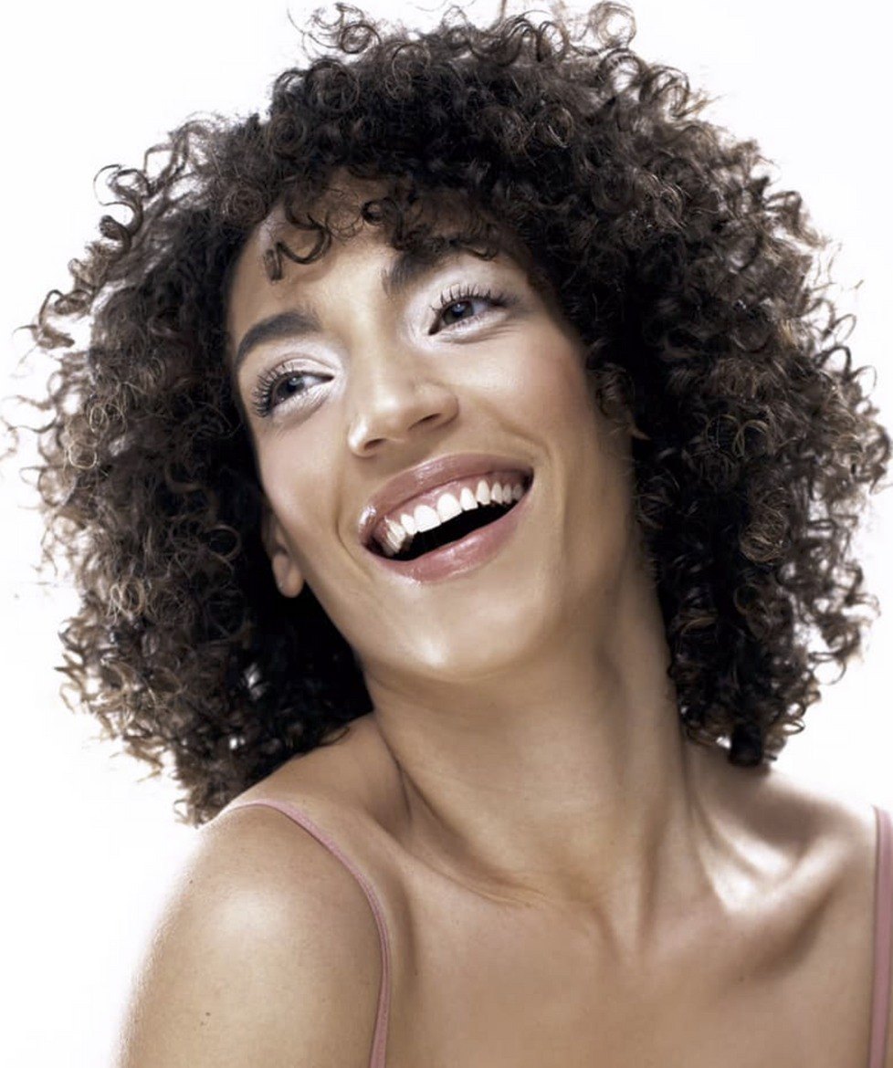 Lachende junge Frau mit voluminösen Locken und trägerlosem rosa Top vor weißem Hintergrund