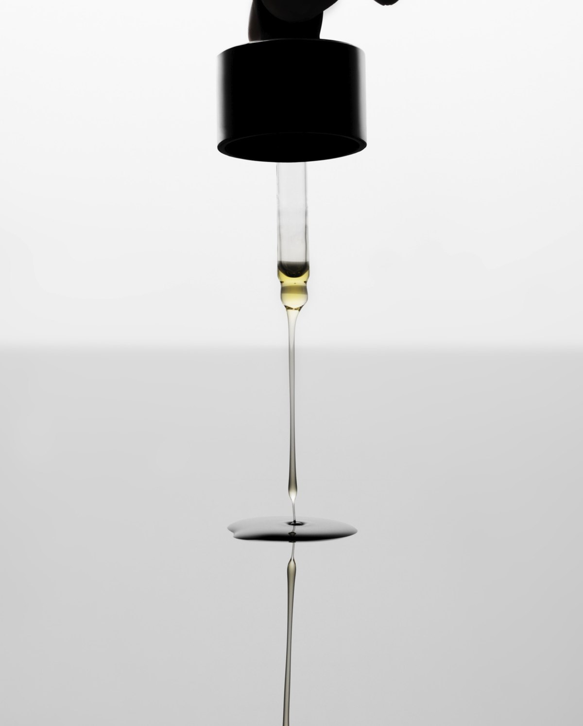 Goldgelber Öltropfen fällt aus einer schwarzen Pipette auf eine spiegelnde Fläche vor hellem Hintergrund