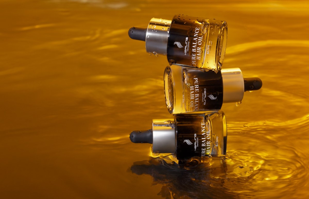 Drei Pure Balance Hair Oil Flaschen liegen gestapelt auf einer goldgelben, bewegten Ölfläche