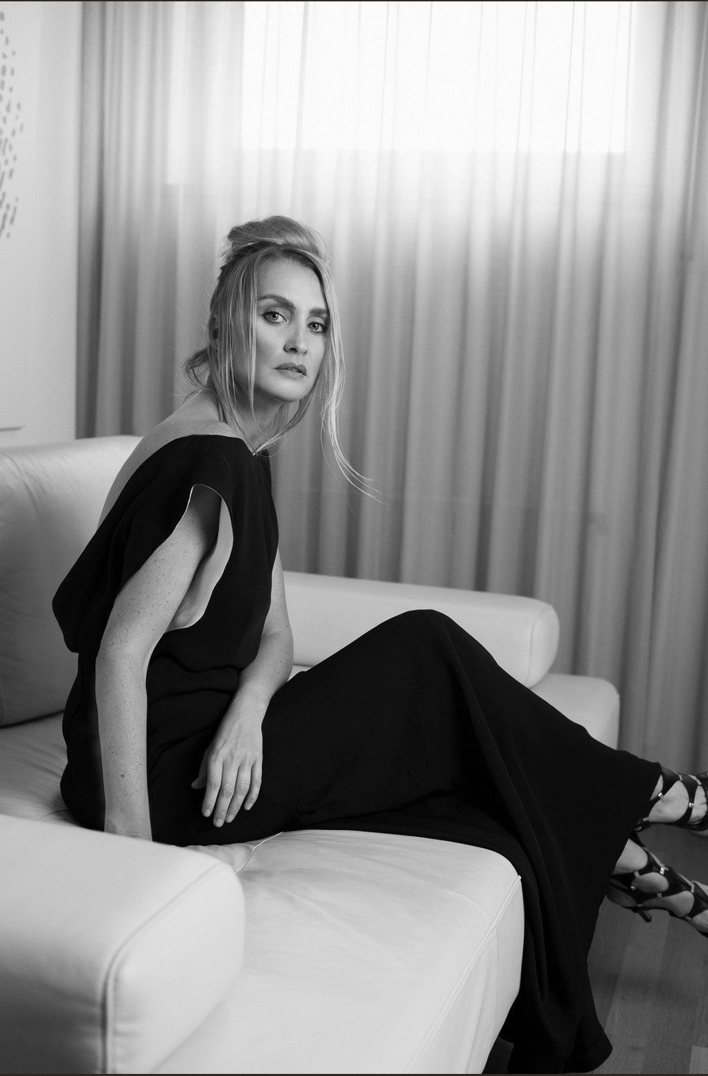 Model im langen schwarzen Kleid auf hellem Sofa vor lichtdurchflutetem Vorhang in Schwarzweiss