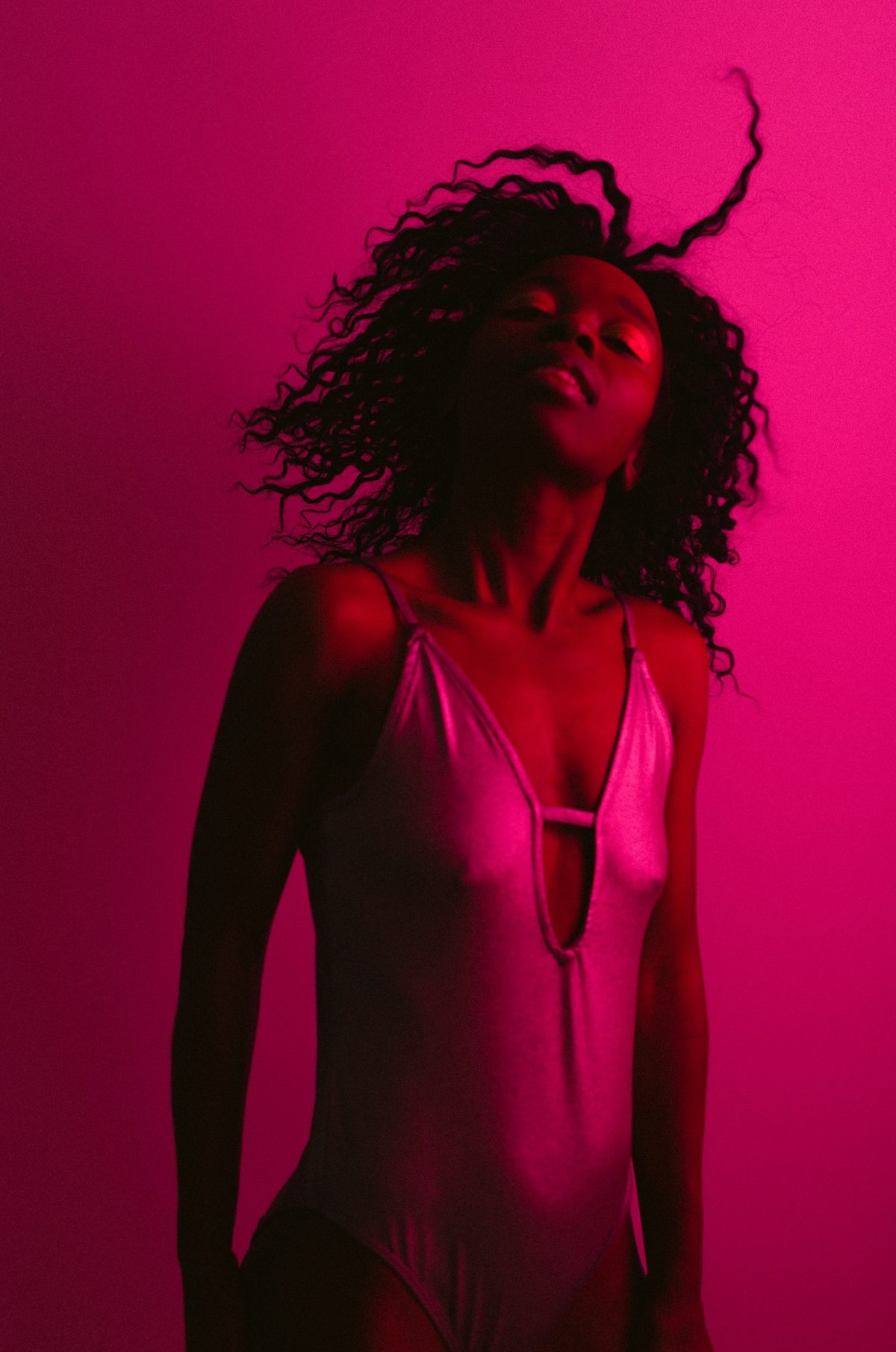 Model im hellen Bodysuit mit fliegenden Locken vor magentafarbenem Hintergrund in farbigem Gel-Licht