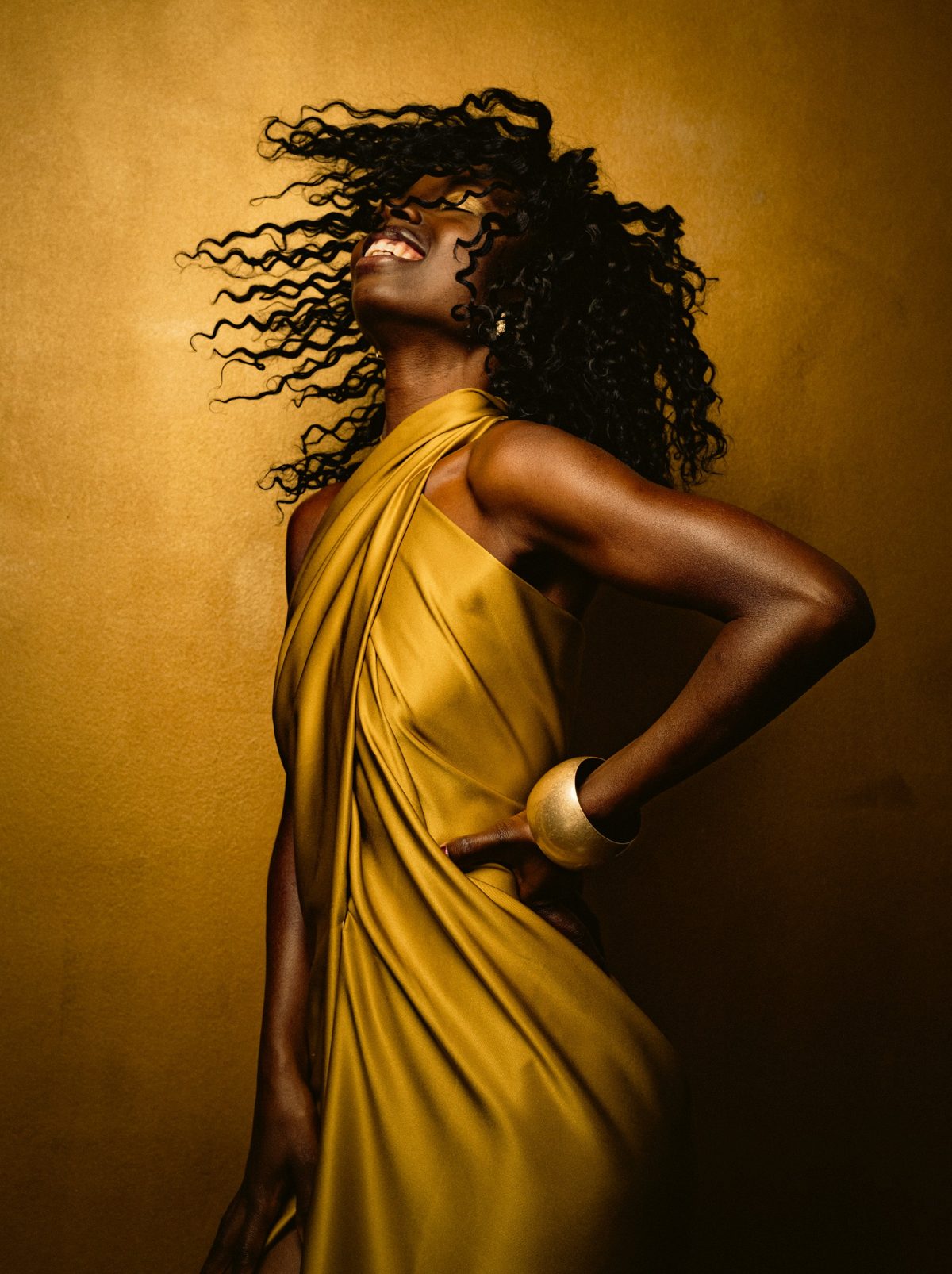 Model in goldgelbem drapiertem Kleid mit fliegendem Haar vor goldbraunem Hintergrund
