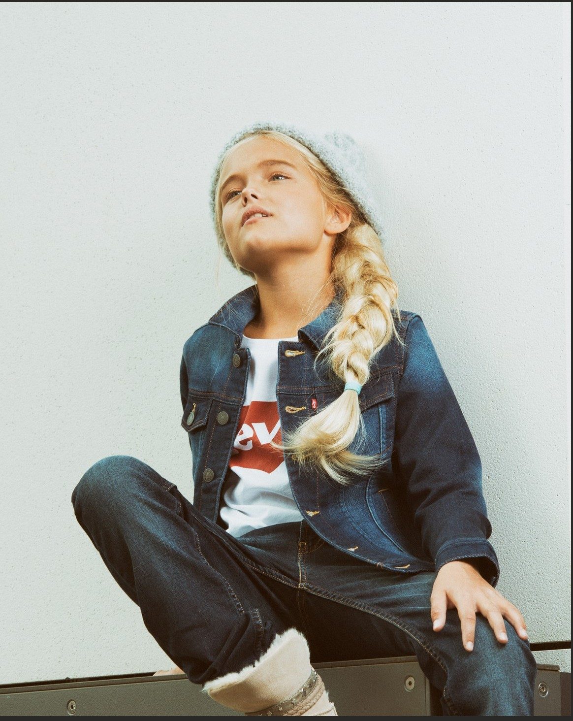 Sitzendes Mädchen mit Strickmütze und Jeansjacke vor heller Wand