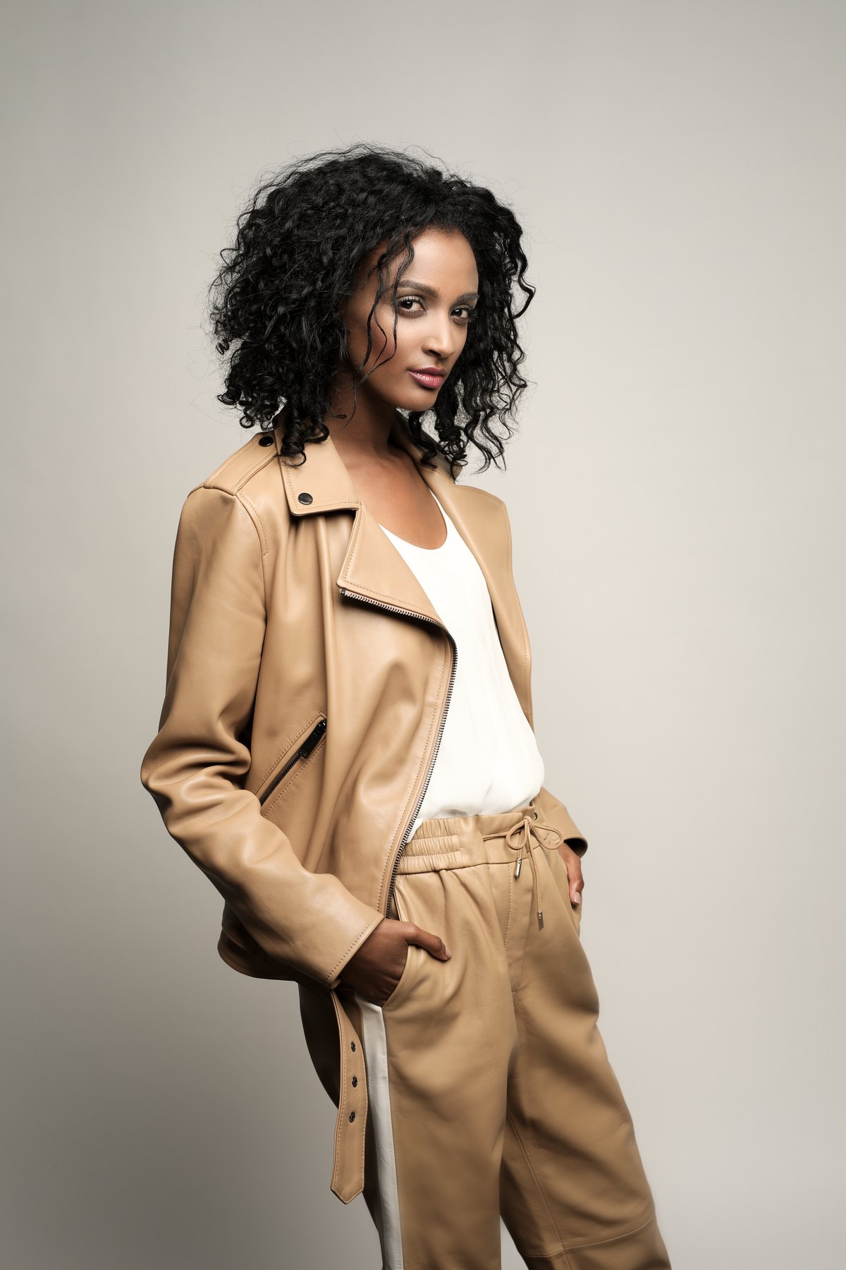 Model in camelfarbener Lederjacke und heller Hose vor grauem Studiohintergrund