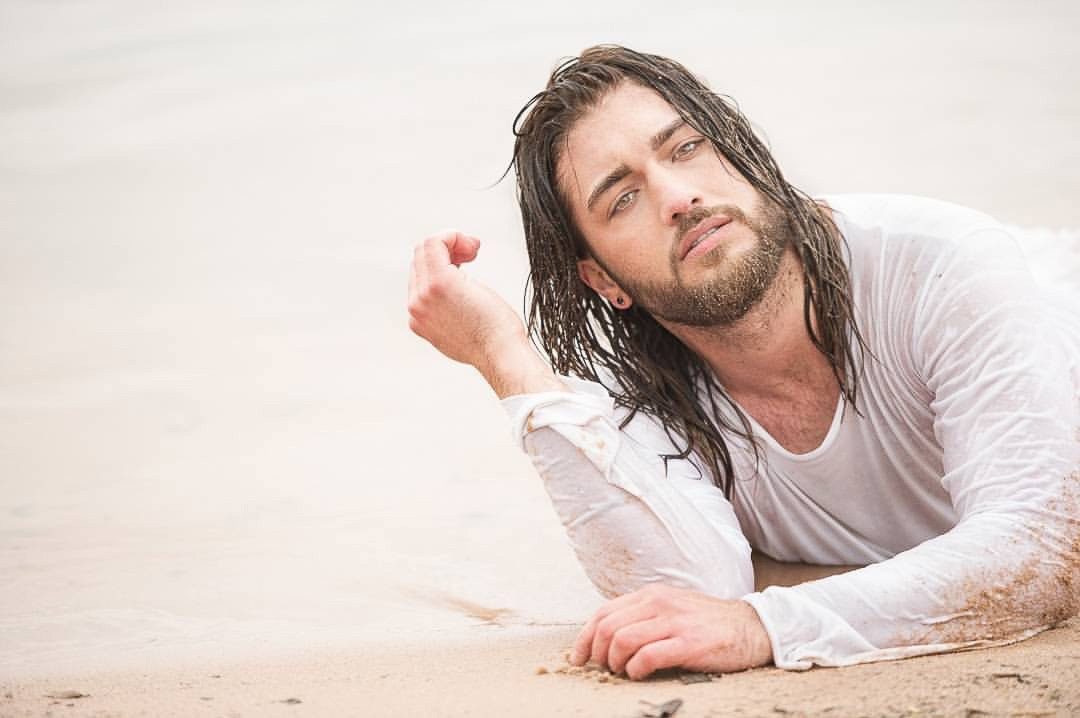 Mann mit nassen langen Haaren und weißem Shirt liegt im hellen Sand am Wasser