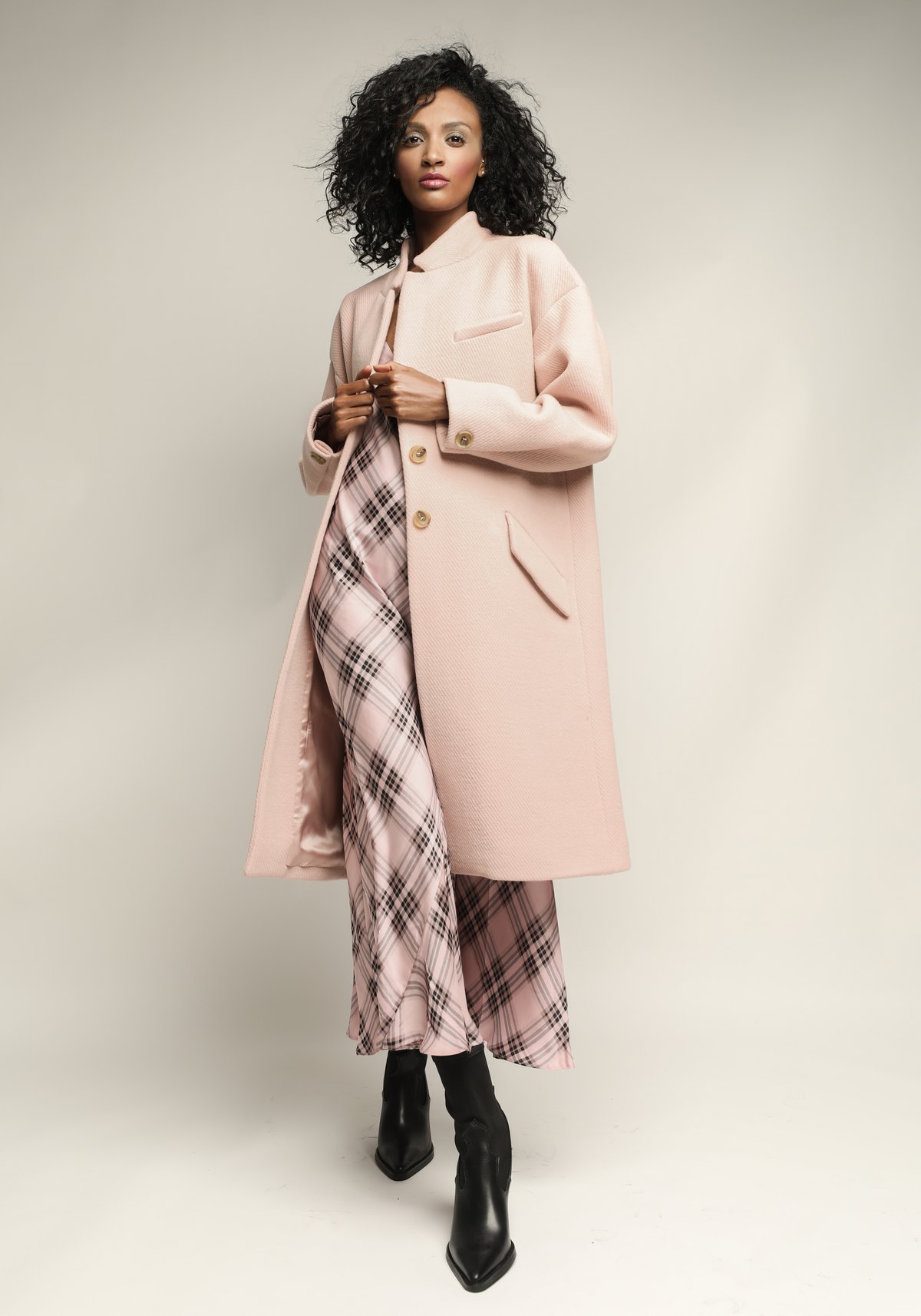Model im rosafarbenen Mantel über kariertem Kleid vor Creme