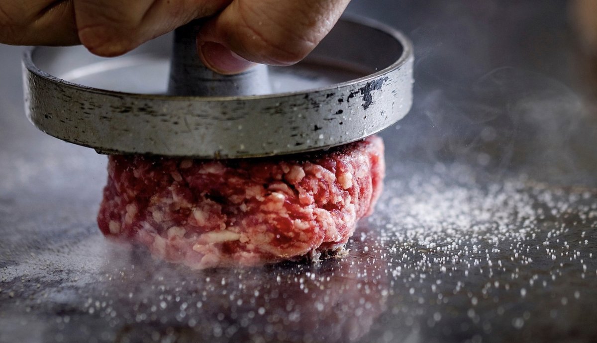 Hand presst ein Burger-Patty aus Rinderhack mit einer Metallpresse auf die heiße Grillplatte