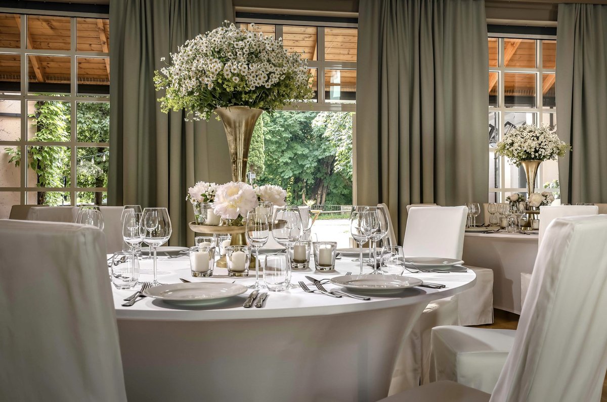 Festlich eingedeckte runde Tafel mit weißen Hussen, Kristallgläsern und hohem Blumenarrangement vor Sprossenfenstern