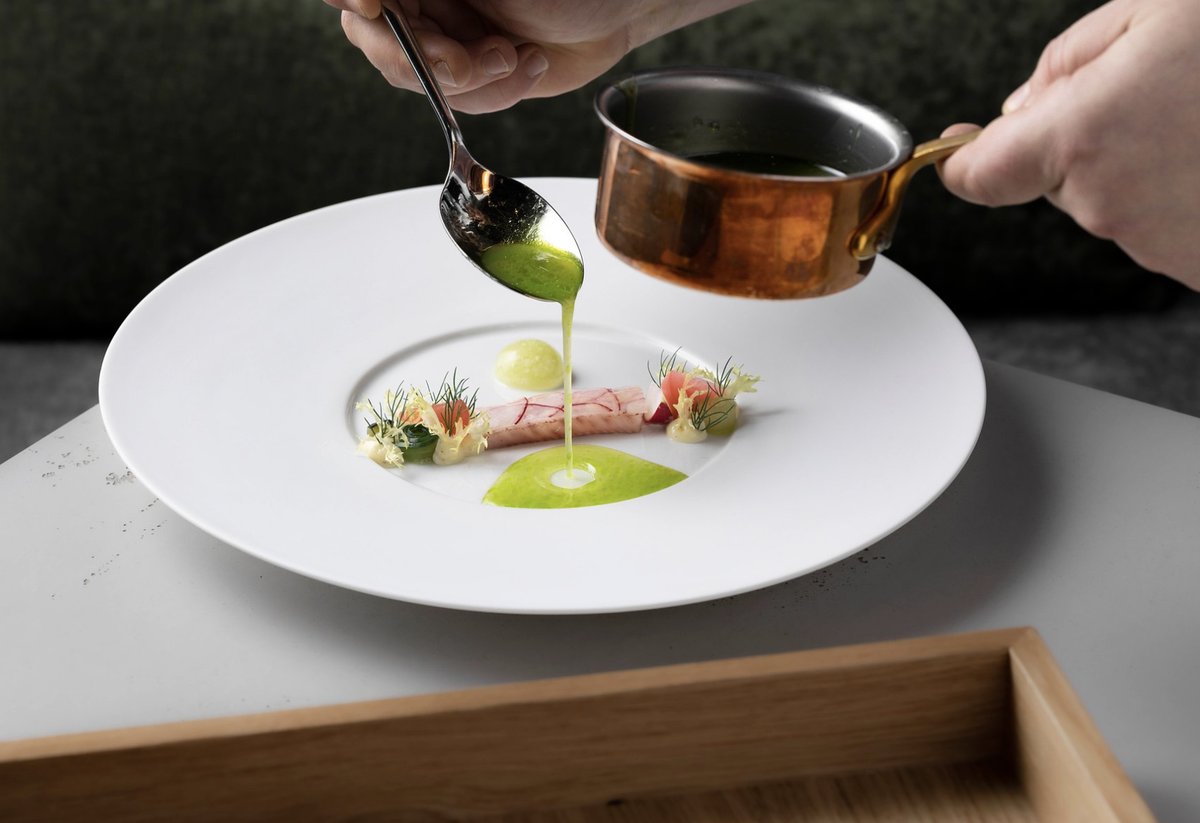 Grüne Sauce läuft vom Löffel auf ein Fischgericht auf weißem Teller, daneben ein Kupfertöpfchen