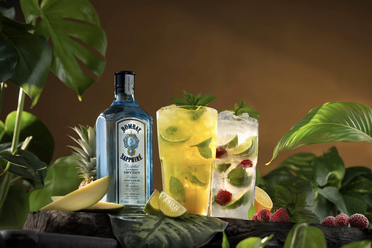 Ginflasche mit zwei Cocktails mit Minze, Limette und Himbeeren zwischen tropischen Pflanzen und Mango