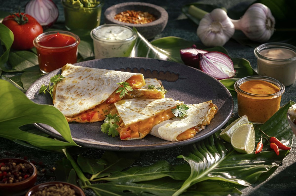 Quesadilla mit geschmolzenem Käse auf dunklem Teller, umgeben von Dips, Zwiebeln, Knoblauch und tropischen Blättern