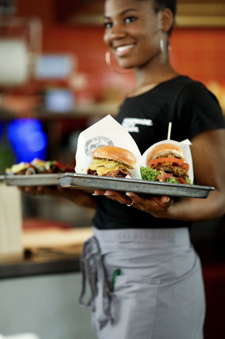 Lächelnde Bedienung trägt ein Tablett mit zwei Burgern von Ruffs Burger