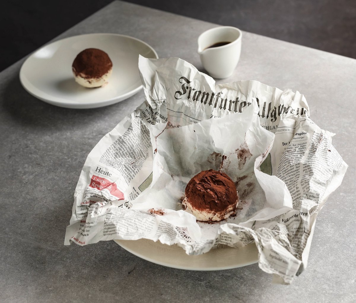 Tiramisu-Kugel mit Kakao in zerknittertem Zeitungspapier, dahinter ein Teller und ein Kännchen Espresso