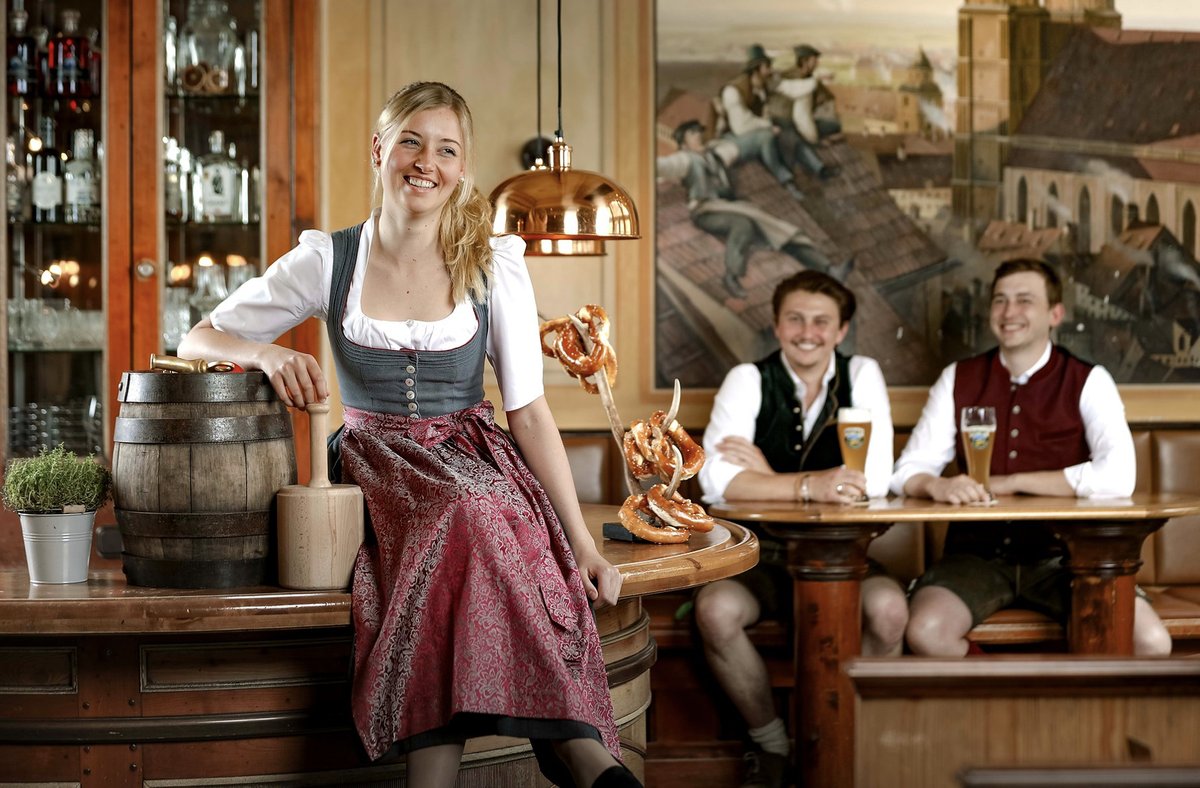 Lachende Bedienung im Dirndl sitzt an der Theke eines Wirtshauses, im Hintergrund zwei Gäste in Tracht mit Weißbier