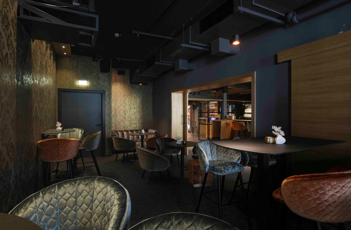 Dunkle Lounge mit Samtsesseln in Petrol und Orange und beleuchteter Bar im Hintergrund