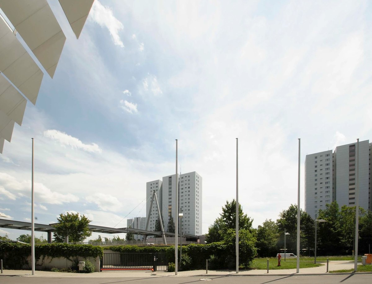 Fahnenmasten vor Wohnhochhäusern und weißem Dachtragwerk unter weitem Wolkenhimmel