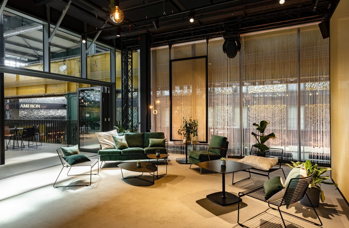 Weiter Blick in einen Lounge-Bereich mit grünen Samtsofas, Glaswand und offener Deckenkonstruktion