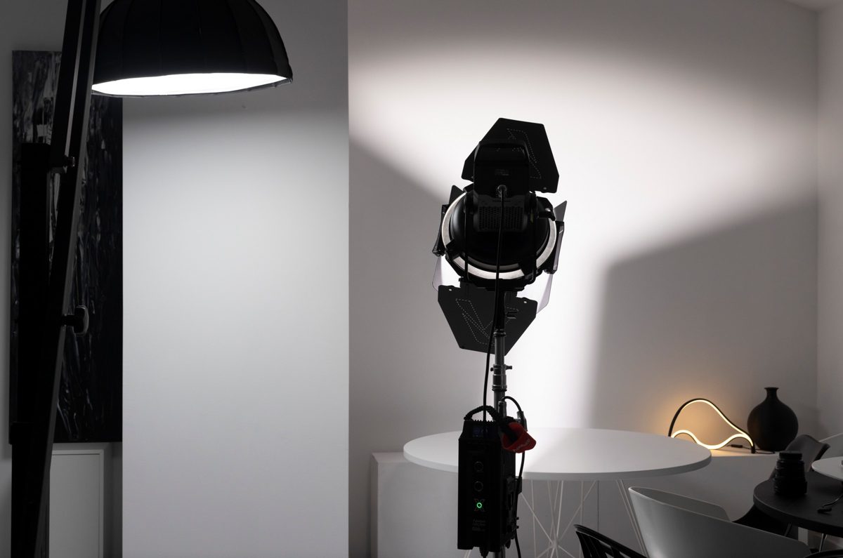 Fotoleuchte mit Flügeltoren auf Stativ leuchtet eine helle Wand an, daneben ein Schirmreflektor