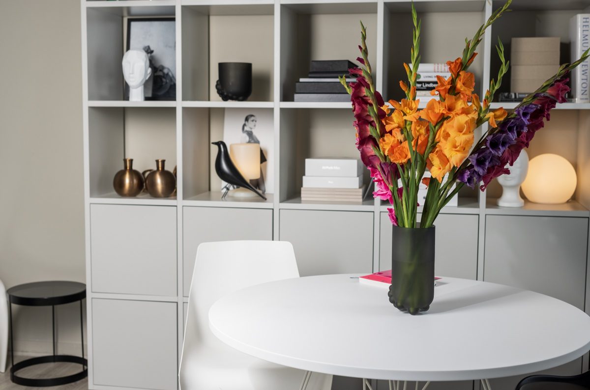 Heller Wohnraum mit weißem Rundtisch und farbenfrohem Gladiolenstrauss vor einem Designregal
