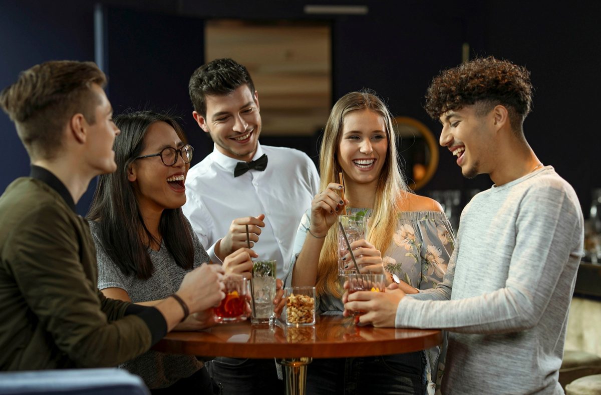 Fünf junge Menschen lachen gemeinsam an einem Bartisch mit Cocktails
