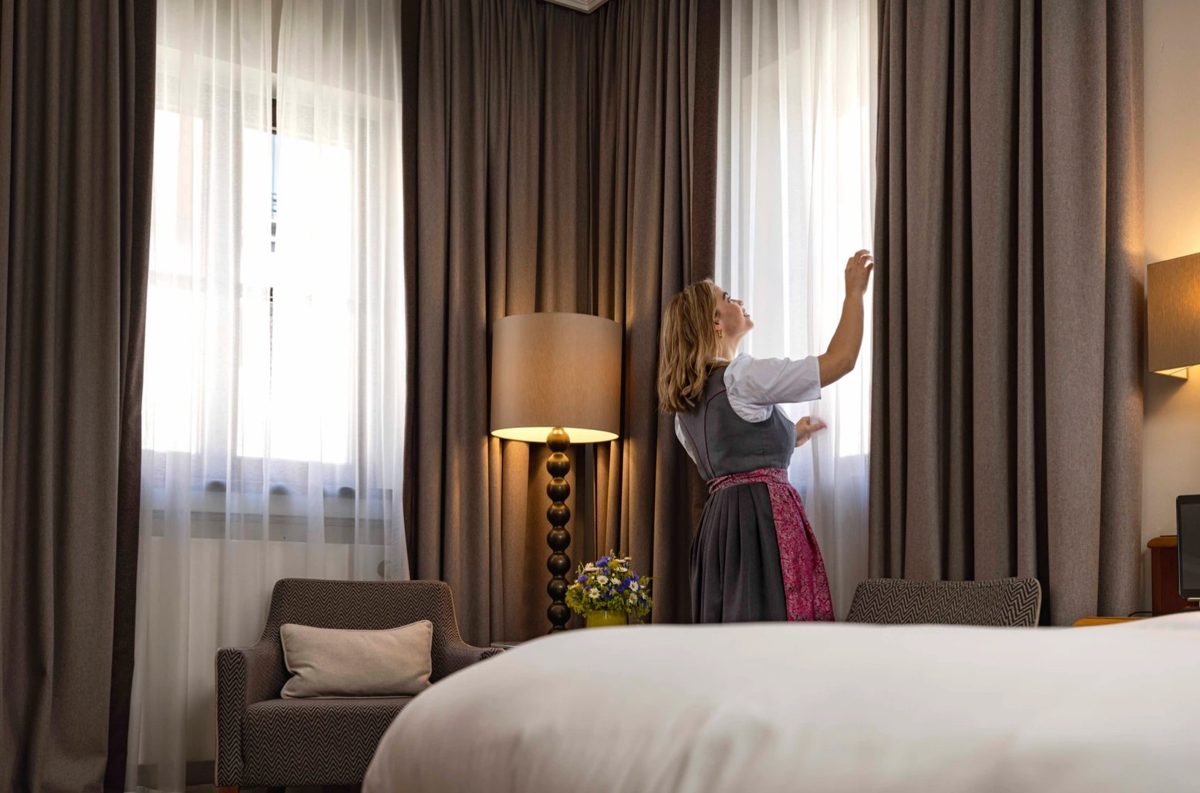Frau in Dirndl öffnet den Vorhang in einem Hotelzimmer mit Bett und Sessel