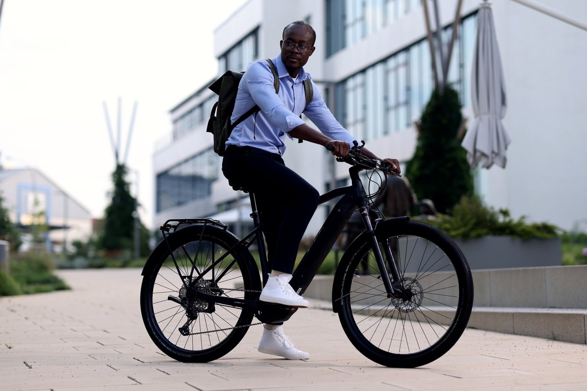 Mann im hellblauen Hemd mit Rucksack sitzt auf einem schwarzen E-Bike vor einem modernen Bürogebäude