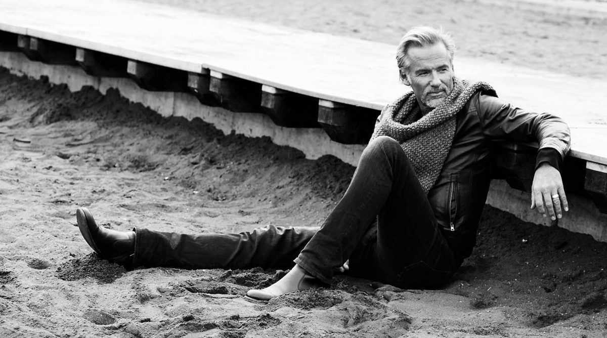 Mann in Lederjacke und Schal sitzt im Sand vor einer Holzplattform, schwarzweiss