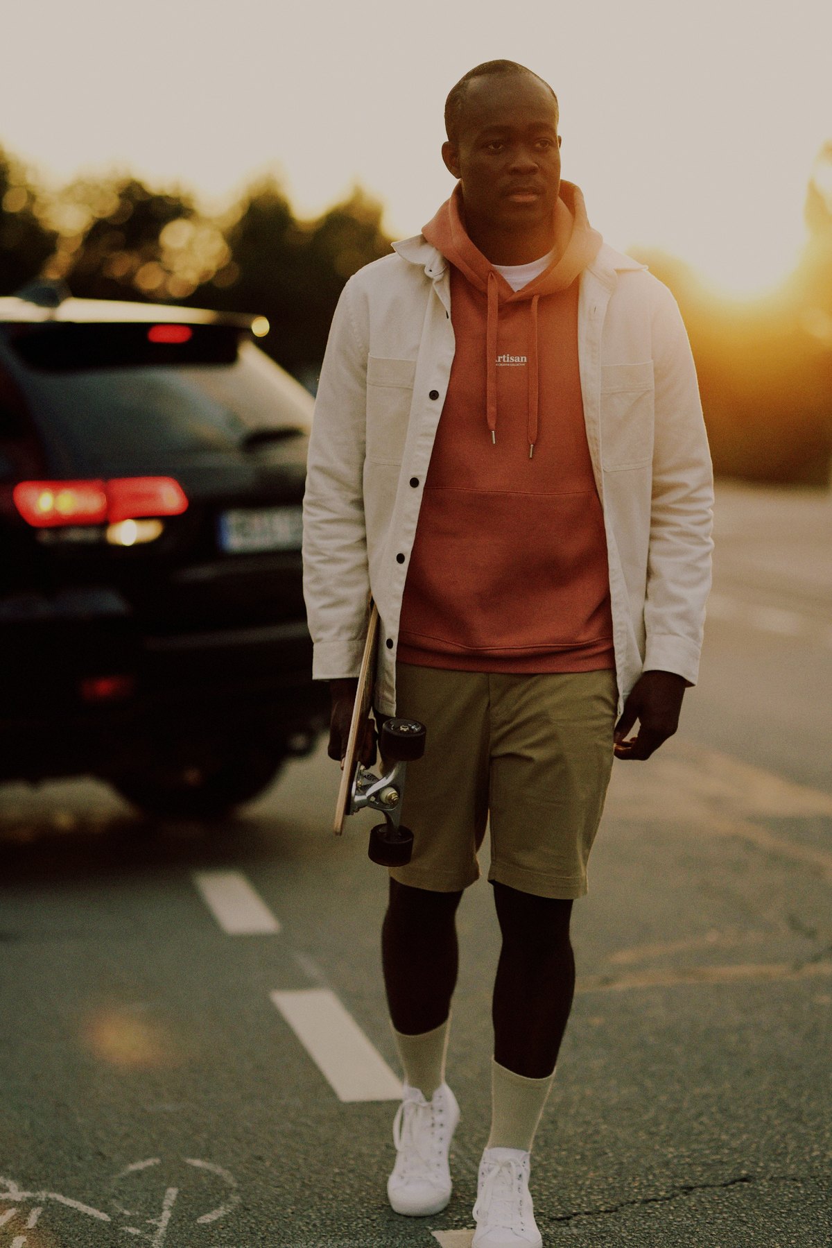 Mann geht mit Longboard in der Hand im Gegenlicht der Abendsonne über eine Straße