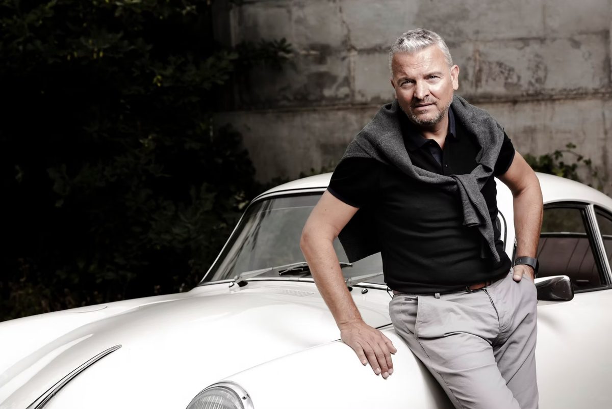 Mann in schwarzem Polo lehnt an einem weißen Oldtimer-Sportwagen vor Betonwand