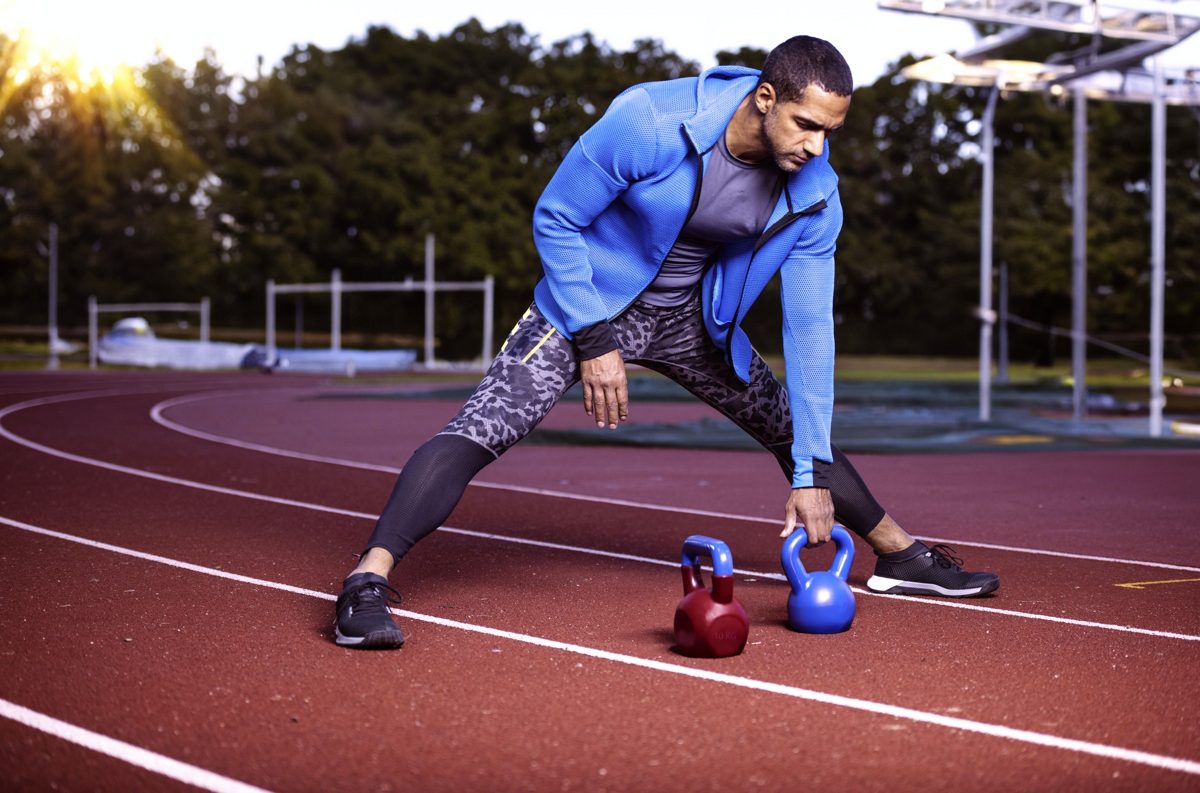 Sportler dehnt sich auf einer roten Laufbahn neben zwei Kettlebells im Gegenlicht