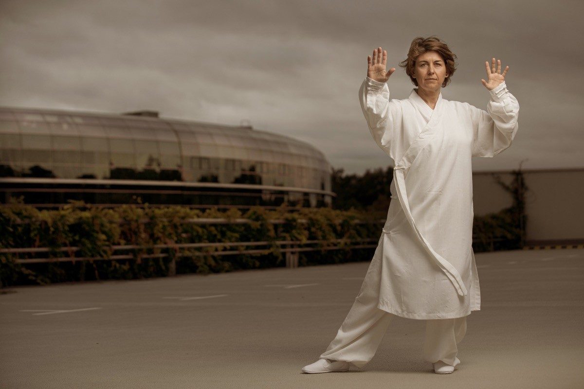 Frau in weißem Gewand in Tai-Chi-Haltung auf einem leeren Parkdeck vor Wolkenhimmel