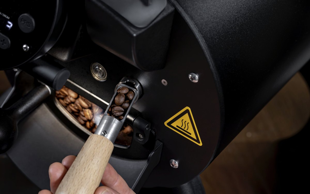 Hand entnimmt mit einem Holzlöffel geröstete Kaffeebohnen aus einem schwarzen Röster