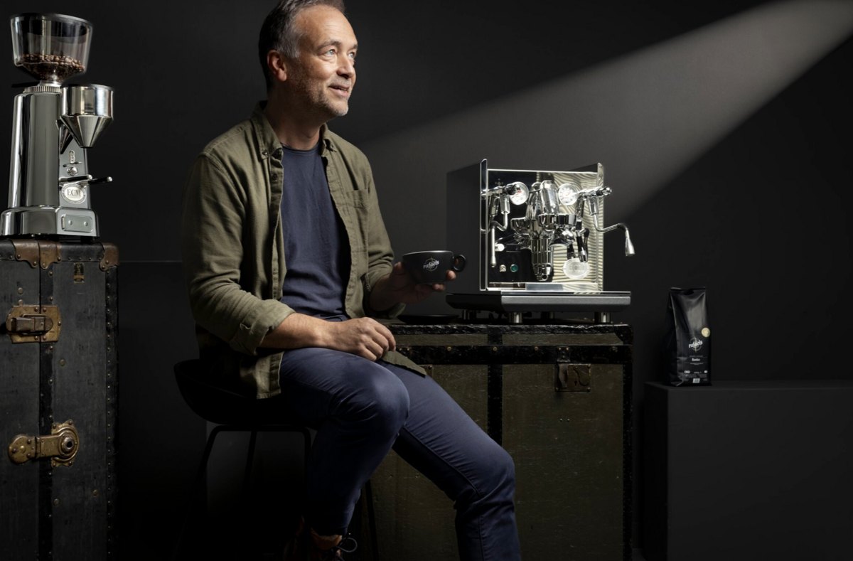 Mann sitzt neben Siebträgermaschine und Mühle und hält eine Kaffeepackung