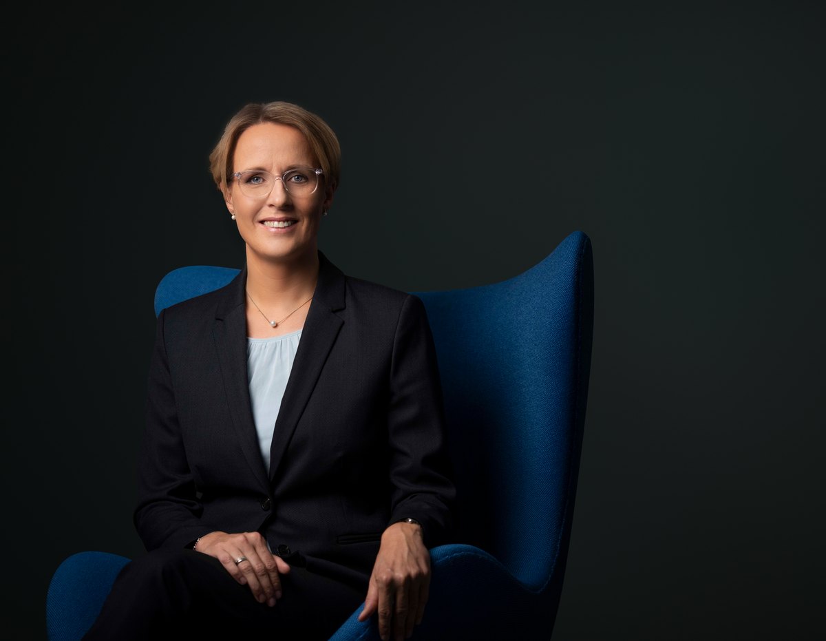 Frau im dunklen Blazer sitzt in blauem Sessel vor dunklem Grund