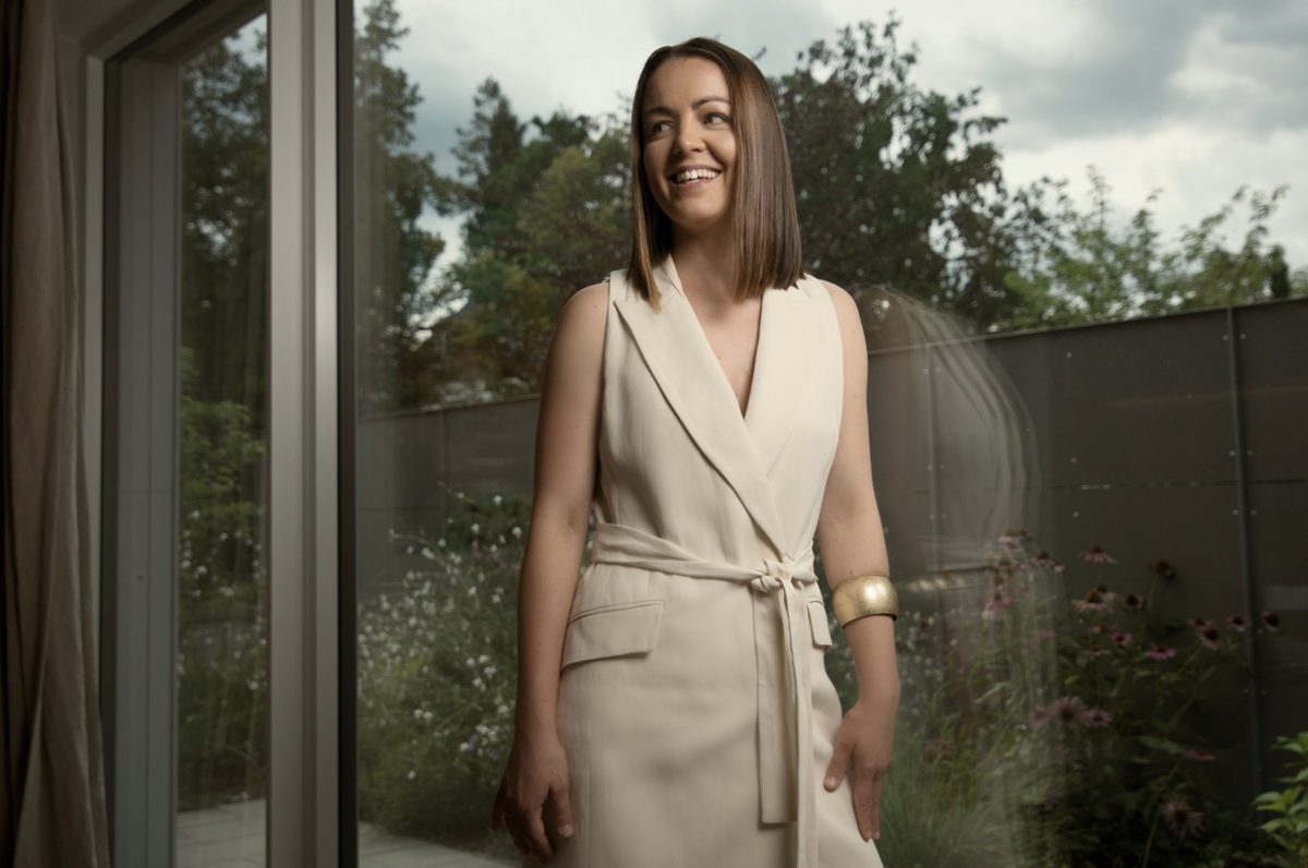 Businessportrait einer lächelnden Frau im cremefarbenen Kleid an einer Glastür mit Gartenblick