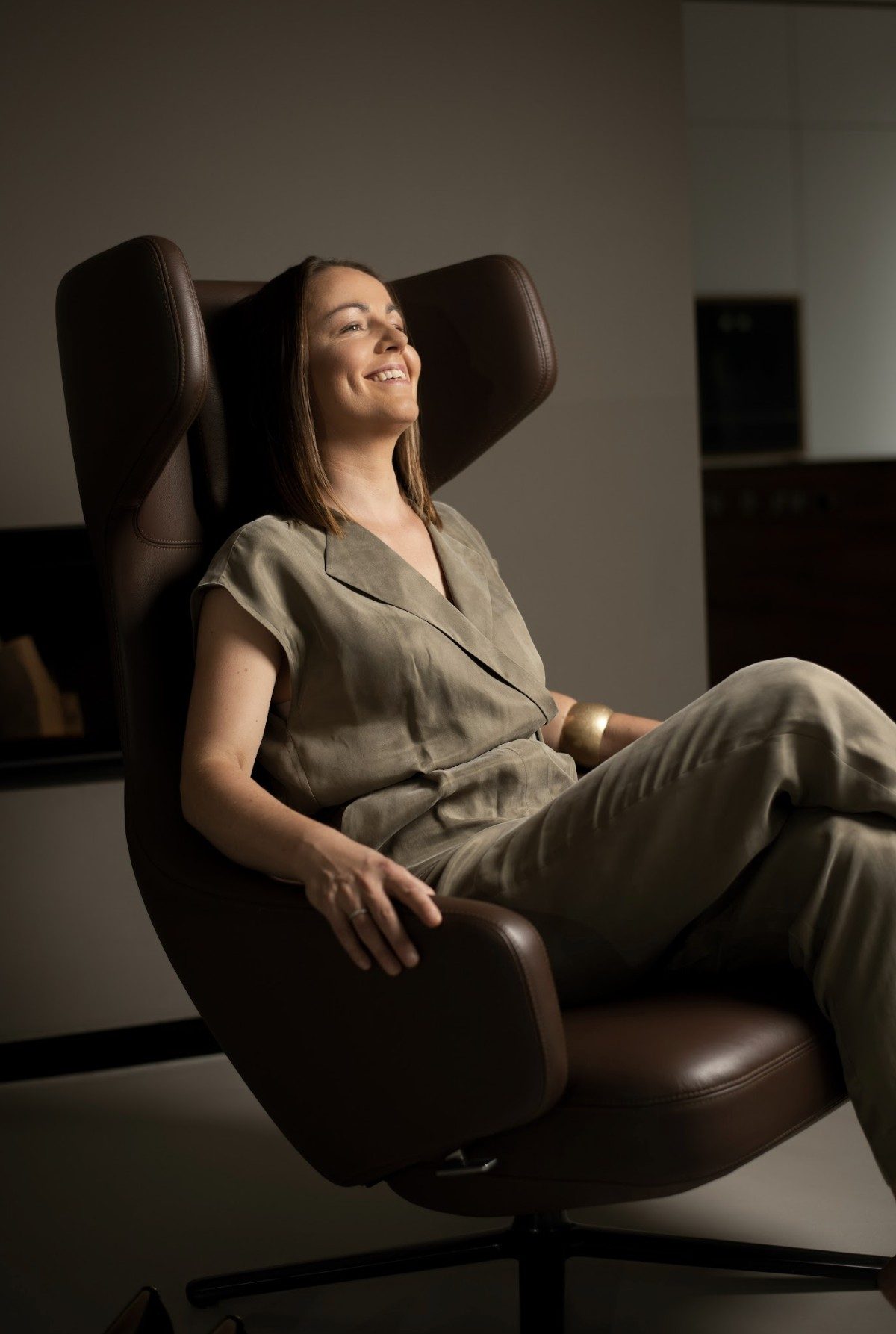 Businessportrait einer lachenden Frau im olivfarbenen Jumpsuit in einem braunen Ledersessel