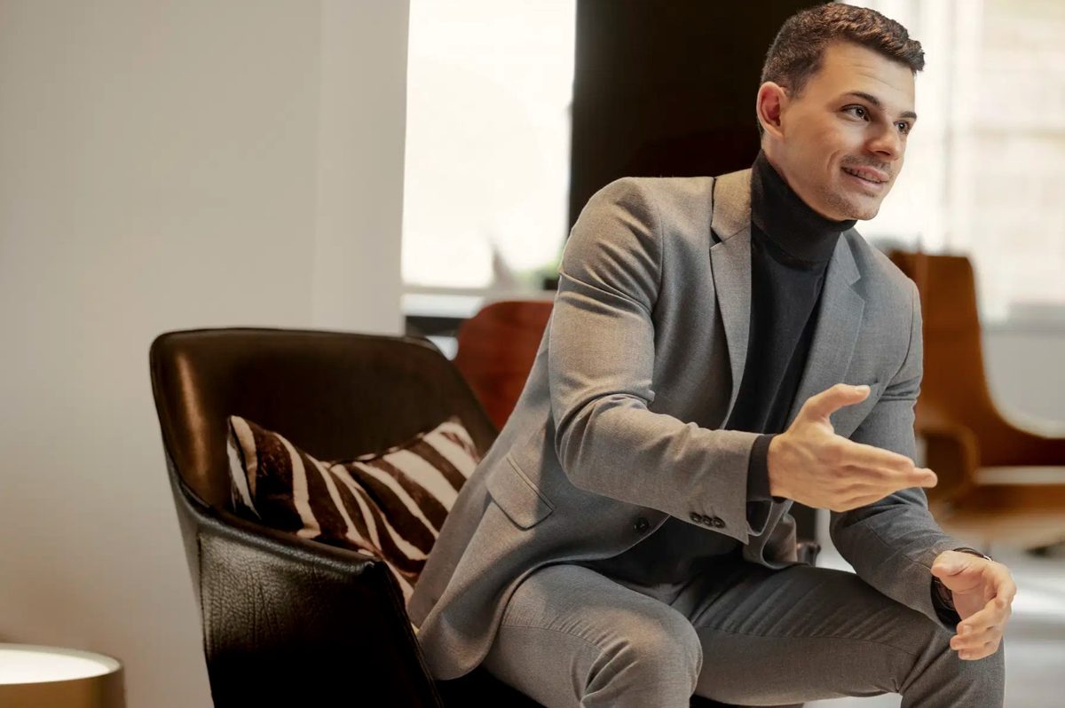 Businessportrait eines gestikulierenden Mannes im grauen Anzug neben einem Ledersessel