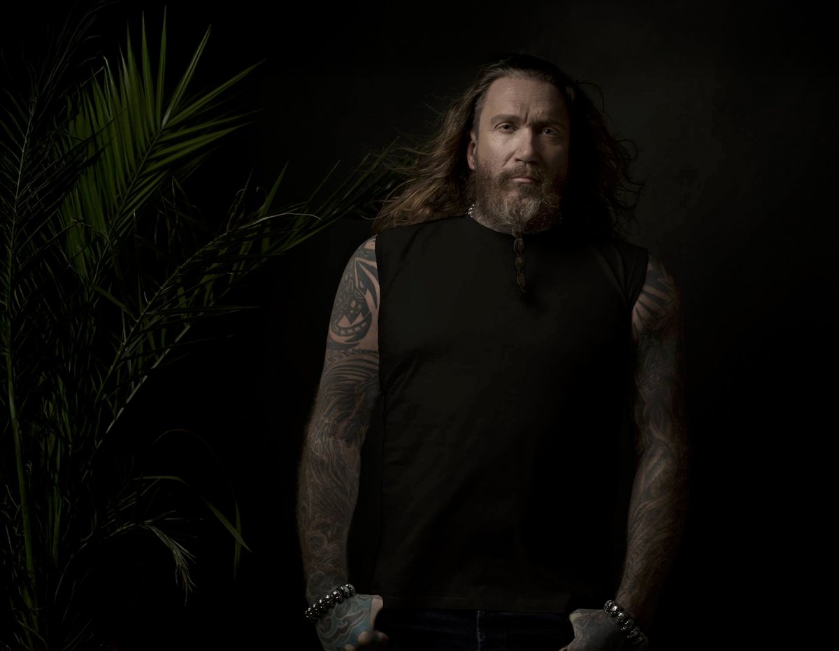 Mann mit langen Haaren und Tattoos vor dunklem Palmenhintergrund