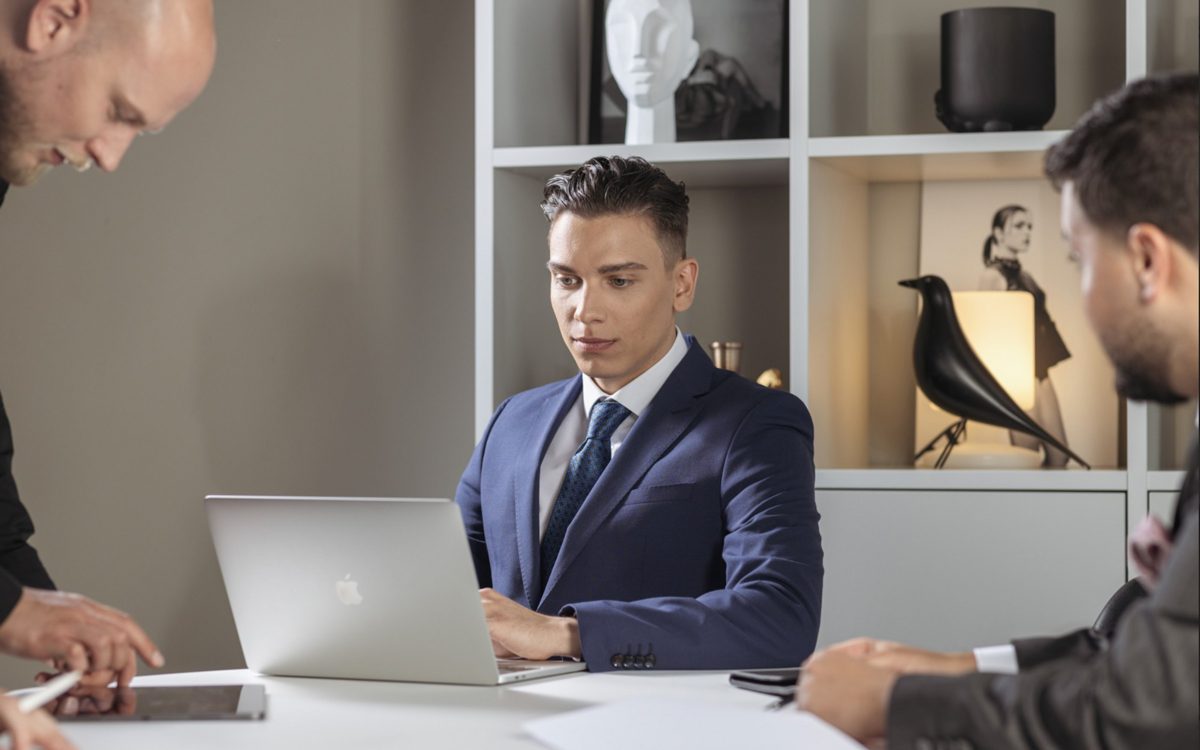 Drei Männer in Anzügen bei einer Besprechung an einem weißen Tisch mit Laptop