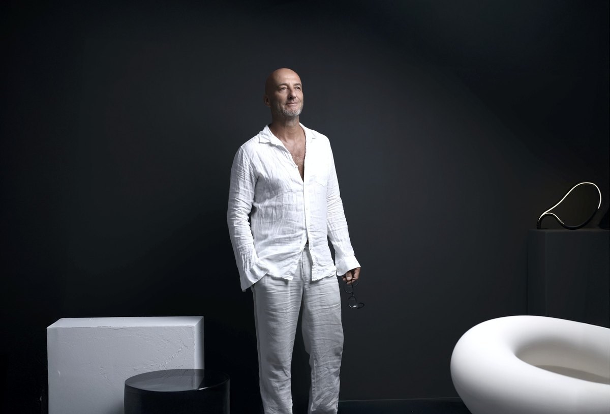 Stehender Mann in weißem Leinenhemd mit Brille in der Hand vor dunkler Wand