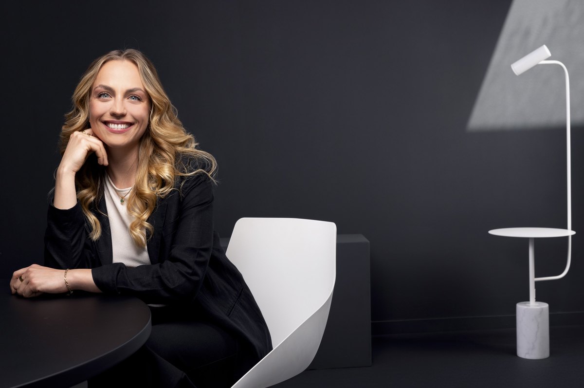 Lächelnde Frau mit blonden Locken sitzt an dunklem Tisch vor weißem Designstuhl