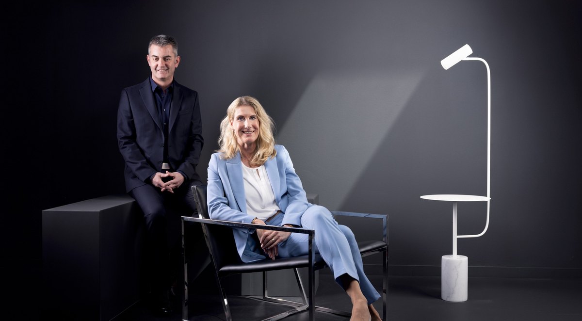 Mann und Frau im Business-Look posieren auf Podest und Lederstuhl vor dunklem Hintergrund