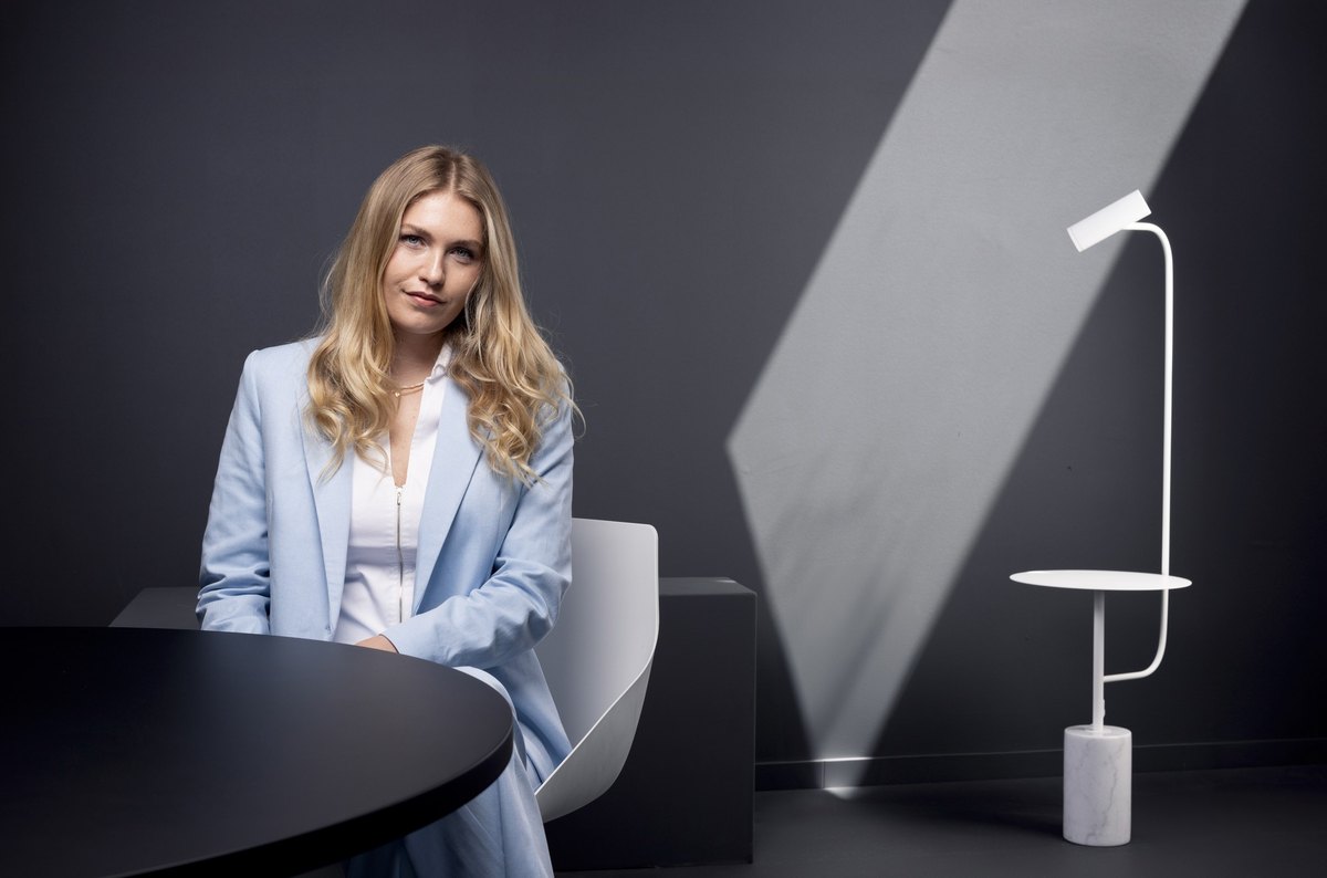 Junge Frau in hellblauem Blazer sitzt an dunklem Tisch vor weißem Designstuhl