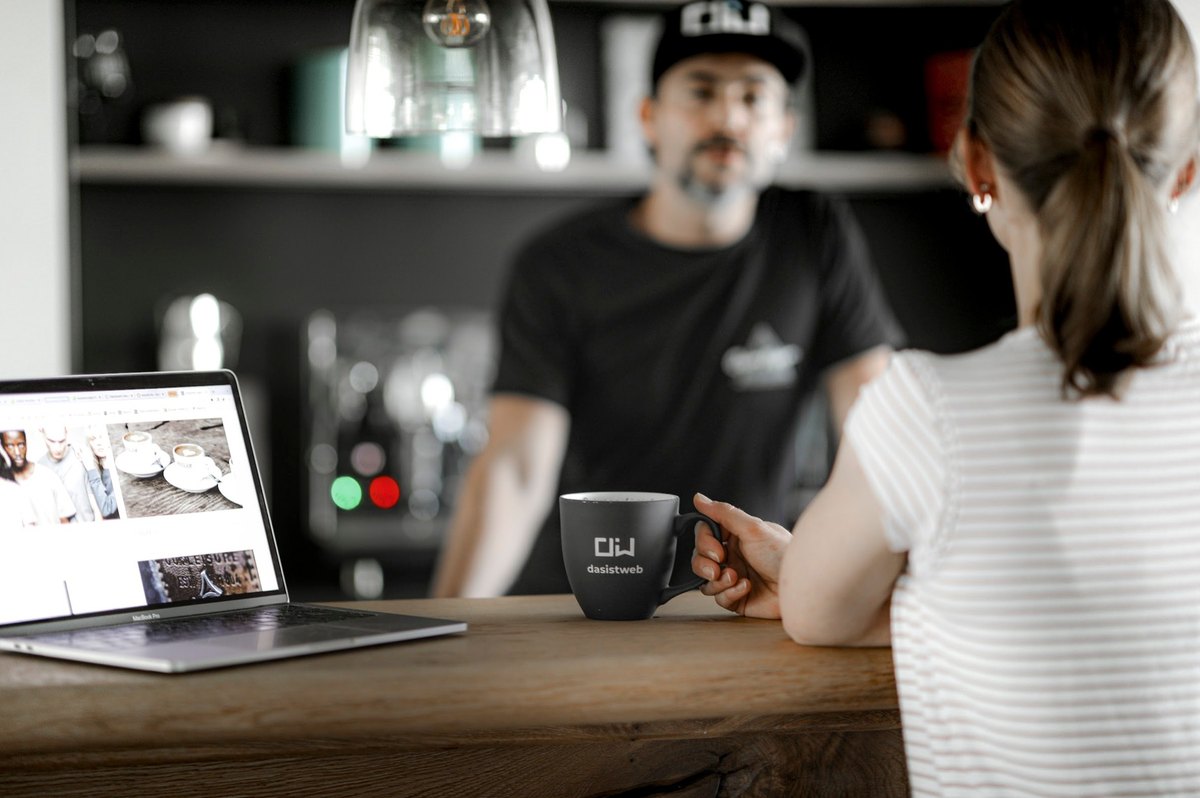 Frau mit Laptop und Kaffeebecher im Gespräch mit Barista an der Theke