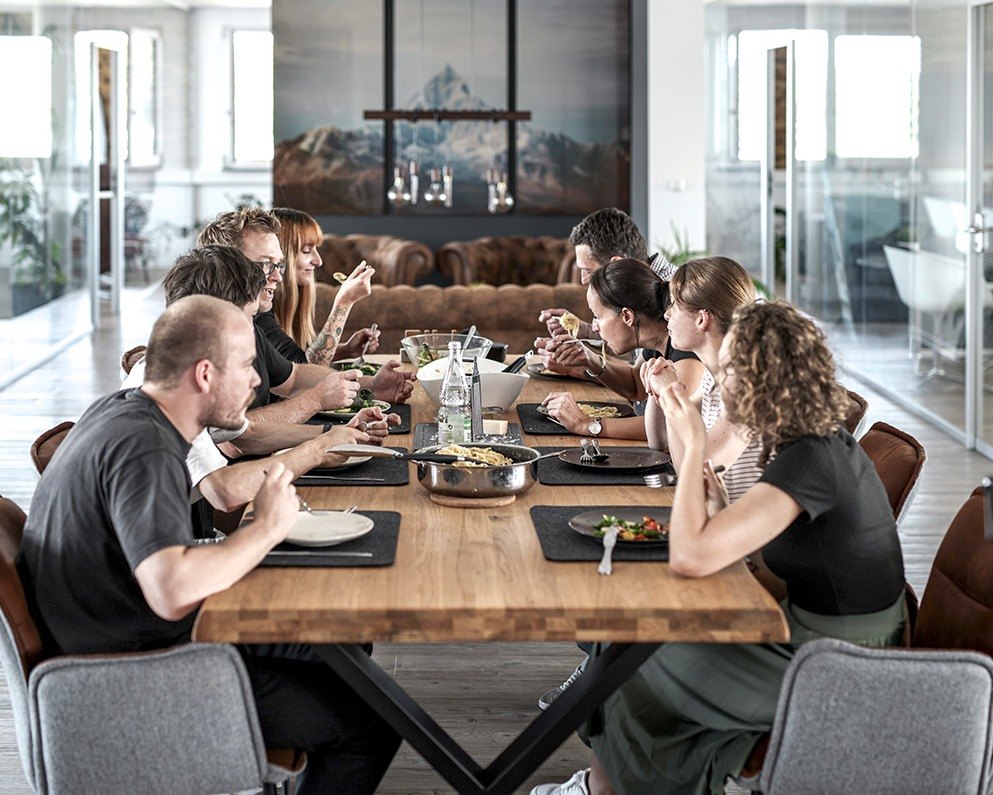 Große Gruppe beim gemeinsamen Mittagessen an langer Holztafel im offenen Büro