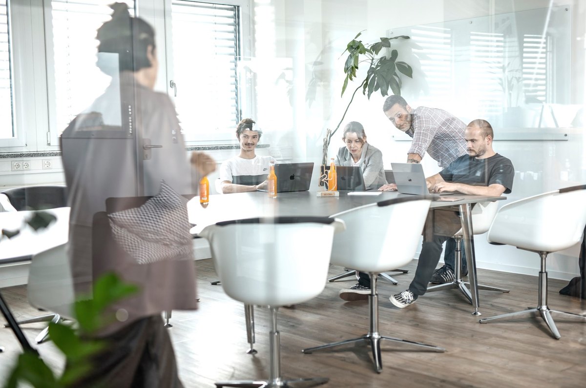 Teambesprechung an rundem Tisch hinter spiegelnder Glaswand im Büro
