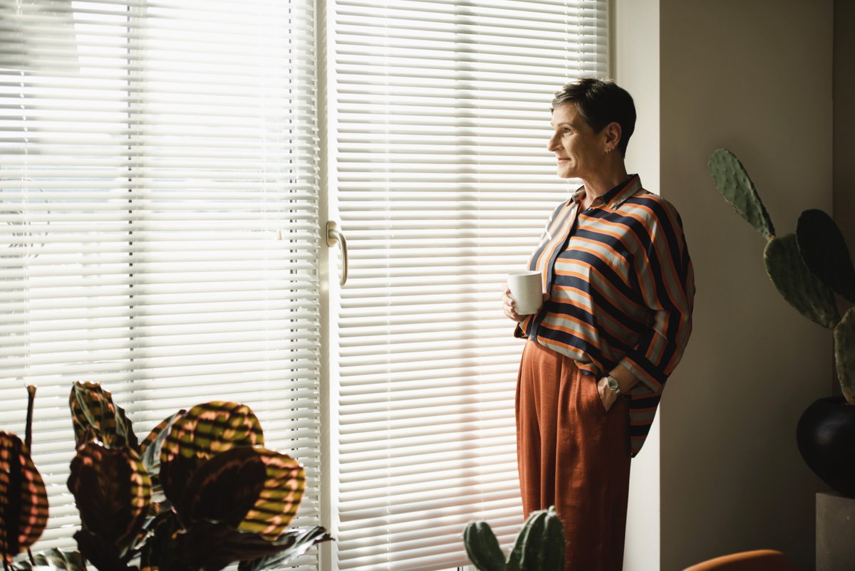 Frau mit Kaffeetasse am Fenster mit Jalousie und Pflanzen