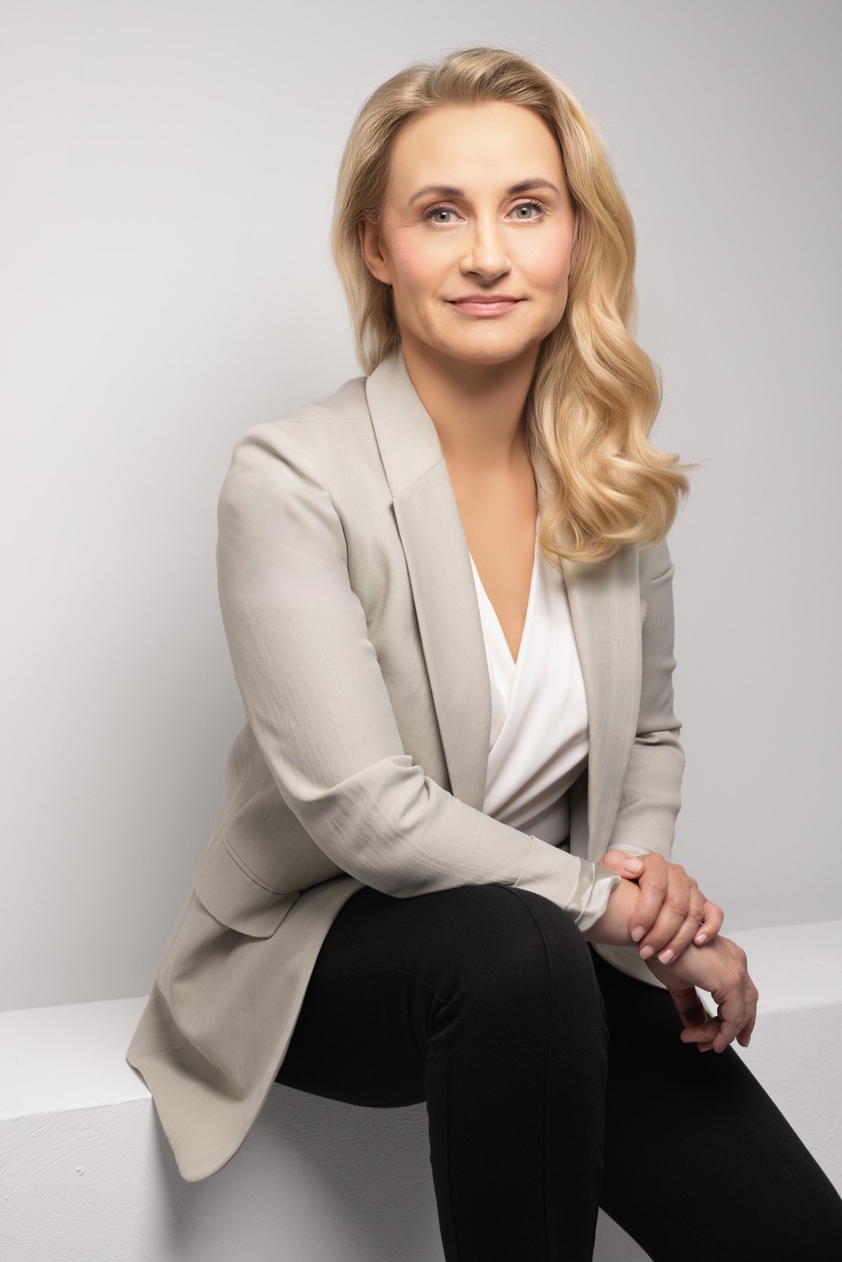 Blonde Frau im hellen Blazer sitzt vor hellgrauem Studiohintergrund